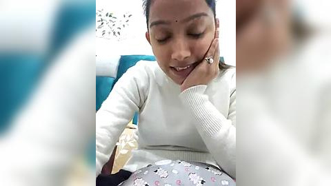 Media: Video of a smiling Indian woman with medium-brown skin, wearing a white sweater, talking on a headset. Background shows a teal chair and a patterned baby blanket.