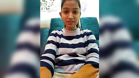 Media: Video of a young girl with medium brown skin, wearing a navy and white striped long-sleeved shirt, seated on a teal sofa, smiling.