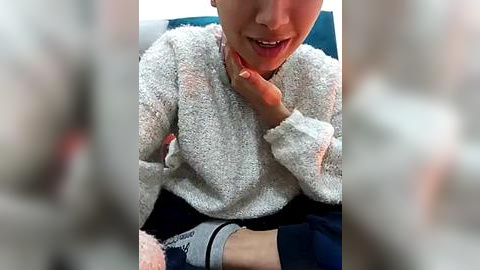 Media: A video of a young woman in a grey sweater, biting her fingernails, seated on a blue sofa, blurred background.