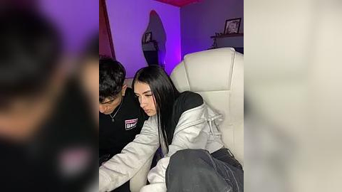 Media: Video of a young woman with long black hair in a gray hoodie, leaning on a man with short dark hair in a black jacket. Background features a dimly lit, modern living room with a purple accent light.