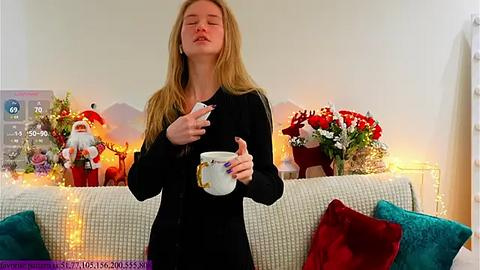 Media: Video of a blonde woman in a black sweater, holding a mug, with a festive background featuring Christmas decorations and a white couch.