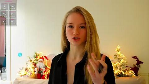 Media: Video of a young blonde woman with fair skin, wearing a black shirt, speaking in a living room with a decorated Christmas tree, a Santa figurine, and lit candles in the background.