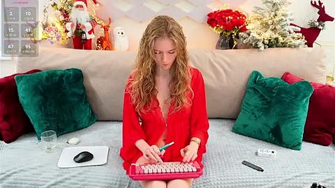 Media: Video of a blonde woman in a red dress, sitting on a beige couch, typing on a pink keyboard. Background features holiday decor including a Christmas tree, reindeer, and festive ornaments.