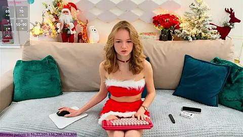 Media: Video of a young, fair-skinned woman with wavy blonde hair, wearing a red Santa-themed outfit, sitting on a beige couch with green pillows, surrounded by Christmas decorations and a keyboard.