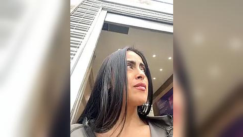 Media: A video of a woman with long black hair, wearing a black leather jacket, looking upward through an open glass door. The background features modern architecture with white blinds and a purple wall.