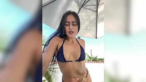 Media: Video of a young woman with long black hair, wearing a dark blue bikini, under an umbrella on a sunny day. She has a tanned complexion and appears relaxed, eyes closed.