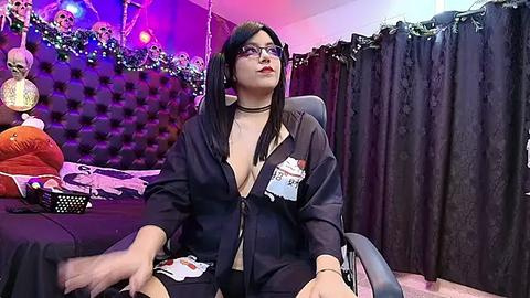 Media: Video of a young, slender Asian woman with long black hair, wearing glasses and a black robe, sitting on a black chair in a dimly lit bedroom with skull decorations, purple lights, and black curtains.