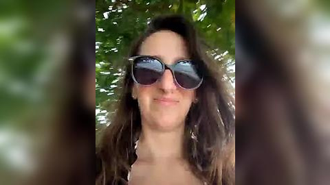 Media: Video of a woman with long brown hair, wearing large, reflective sunglasses, standing outdoors among green foliage. Her facial expression is neutral, and she appears to be smiling slightly.