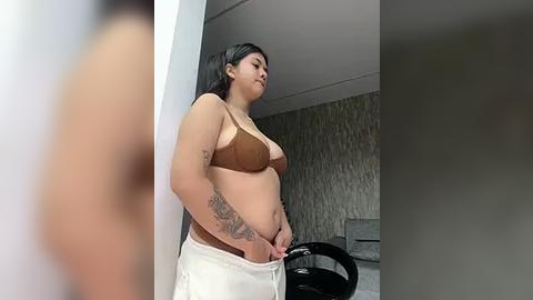 Media: Video of a plus-size Latina woman with medium skin tone, long black hair, and tattoos on her arms, wearing a brown bra and white towel, standing in a bathroom with a tiled wall and a black chair.