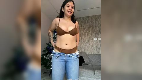 Media: Video of a plus-size woman with medium skin tone and dark hair, wearing a brown bra and unbuttoned jeans, standing in a living room with a grey sofa and a potted plant.