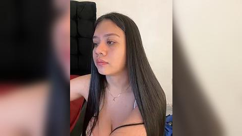 Media: Video of a young Latina woman with long black hair and light skin, wearing a black spaghetti-strap top, sitting on a red couch, against a beige wall.