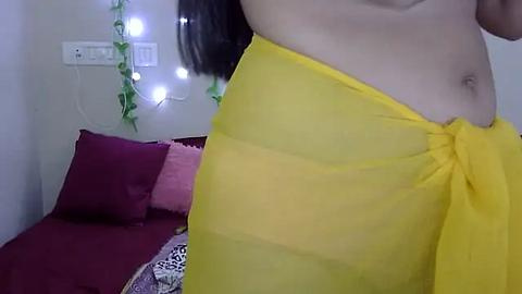 Media: Video of a light-skinned woman's midsection, wearing a yellow sheer wrap, standing next to a bed with maroon and pink bedding, and green fairy lights in the background.