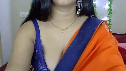 Media: Video of a woman with long black hair wearing a blue blouse and an orange sari with a gold necklace. Background features a white wall, electrical outlet, and a green plant.