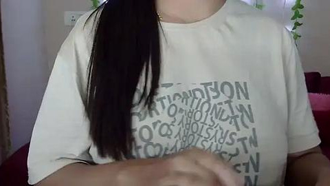 Media: Video of a woman with long, straight black hair, wearing a white t-shirt with \"TRIANGULAR\" written in bold gray letters. Background features a light-colored wall, a power outlet, and a glimpse of a red couch.