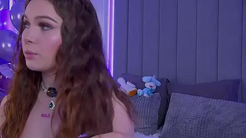 Media: Video of a young, fair-skinned woman with long, wavy brown hair, wearing a black necklace, standing in a dimly lit room with purple lighting, holding a stuffed animal.