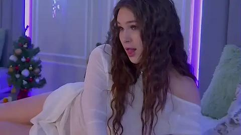 Media: Video of a young woman with long, curly brown hair, wearing a white off-shoulder dress, lying on a bed, with a lit Christmas tree in the background.