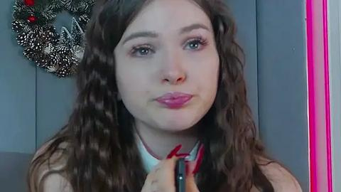 Media: Video of a young woman with long, wavy brown hair, wearing red lipstick, applying makeup with a black brush, in a room with a gray wall, a pink mirror, and a festive wreath.