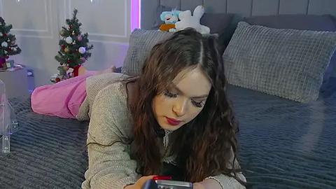 Media: A video of a young woman with long brown hair, red lipstick, and a beige sweater, lying on a grey quilted bed, reading a tablet. The room is decorated with Christmas decor, including a small tree and stuffed toys.