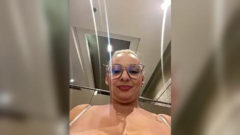 Media: Video of a blonde woman with fair skin, wearing glasses and a white tank top, standing in an elevator with modern, minimalist decor.