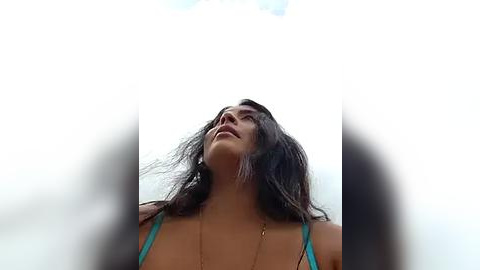 Media: Video of a young woman with long, dark hair, wearing a turquoise bikini top, gazing upwards with a serene expression, against a bright, clear sky.