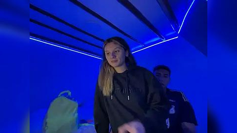 Media: Video of a young woman with long blonde hair in a black hoodie walking down a dimly lit, blue-lit hallway, with two men behind her.