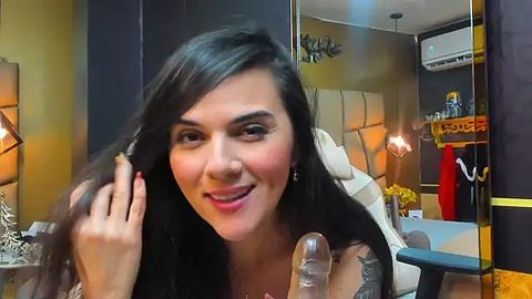 Media: Video of a smiling woman with long dark hair, wearing a white top, in a cozy, dimly lit room with modern decor, including a mirror and lamp.