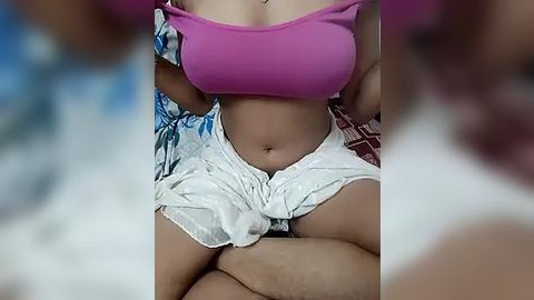 Media: Video of a light-skinned woman in a tight pink crop top and white shorts, seated on a bed with a blurred background.