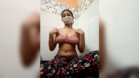 Media: A video of a young woman with medium brown skin, wearing a pink bra, floral pants, and a face mask, sitting in a tiled bathroom with floral-patterned wallpaper.