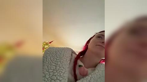 Media: A video featuring a fair-skinned woman with red hair, wearing a light-colored sweater, lying in a bed with a soft, textured blanket. The background is out of focus, creating a warm, cozy atmosphere.