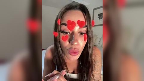 Media: Video of a young woman with long brown hair, wearing a white top, with red heart emojis covering her face. She is indoors, with blurred background showing a bed and wall art.