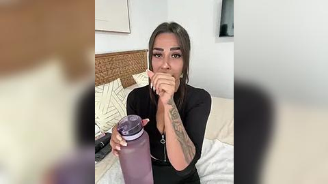 Media: Video of a Latina woman with long dark hair, wearing a black top, holding a pink water bottle, seated on a bed with white sheets and wicker headboard in a modern bedroom.