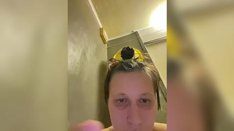 Media: Video of a woman with light skin and brown hair, wearing a yellow headband and a black beanie, standing in a beige bathroom with a shower curtain.