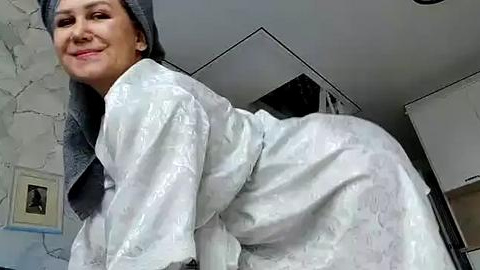 Media: Video of a woman in a white satin robe with a gray towel on her head, leaning forward in a bathroom with light blue walls and a white cabinet.