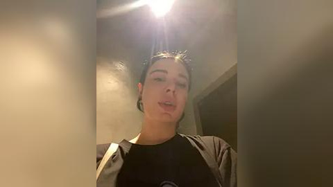 Media: A video of a young woman with fair skin, short dark hair, and wearing a black t-shirt under a grey jacket, standing in a dimly lit, narrow hallway with a bright light above.