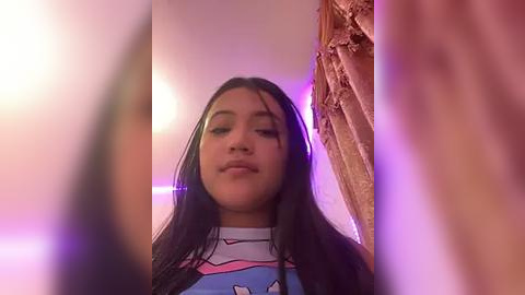 Media: Video of a young Latina woman with long black hair, wearing a colorful graphic t-shirt, standing in a dimly lit room with pink and purple lights.