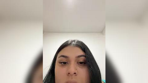 Media: Video of a young woman with long, straight black hair, tan skin, and light makeup, including subtle eyeshadow and eyeliner, captured from below, showing her facial features and nose ring. Background is a plain white ceiling.