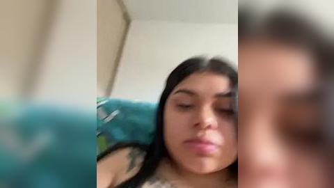 Media: A blurry video shows a young woman with long black hair, resting her head on a teal cushion, eyes closed. The background is out of focus, with a beige wall and indistinct objects.