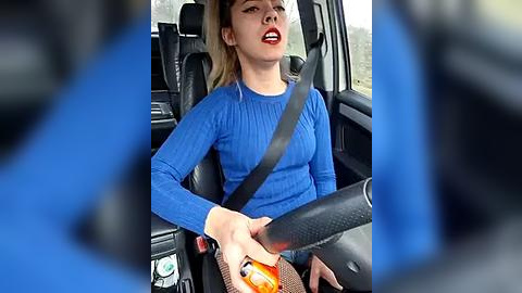 Media: Video of a blonde woman in a blue sweater, driving a car with a safety belt on. She has a serious expression, holding a bottle of pills.