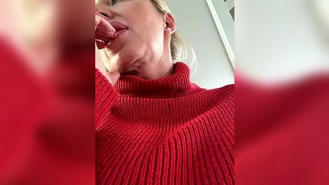 Media: Video of a blonde woman in a red sweater licking her fingers, taken from a low angle, with blurred red objects in the foreground.