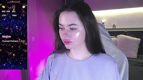 Media: Video of a young woman with long dark hair, wearing a light purple shirt, sitting in a dimly lit bedroom with a TV displaying a customizable filter and a bed with fluffy pillows in the background.