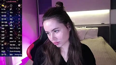 Media: Video of a young woman with long brown hair in a top bun, wearing a black top, seated in a black and red gaming chair in a modern, dimly lit bedroom with a bed and a large screen showing a game interface.