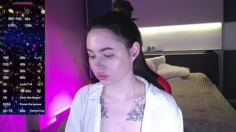 Media: Video of a young Asian woman with fair skin, black hair tied in a high ponytail, wearing a white shirt exposing a floral tattoo on her chest. Background features a modern bedroom with a bed and a starry night sky display.