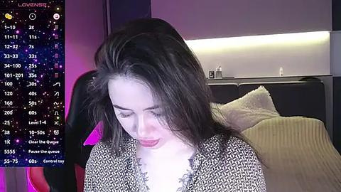 Media: Video of a young woman with long dark hair, wearing a patterned top, sitting on a bed with a yellow pillow. Background includes a digital star chart and dim lighting.