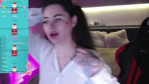 Media: Video of a woman with long dark hair, wearing a white shirt, looking relaxed, sitting in a gaming chair. Background includes a bed with pillows.