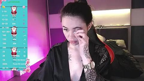 Media: A video of an East Asian woman with dark hair in a bun, wearing a black kimono with lace, sitting on a bed. She's looking at her phone with a thoughtful expression. The background includes a pink and purple ambient light.