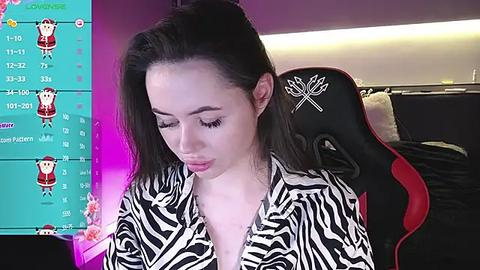 Media: A video of a young woman with fair skin, long dark hair, and large eyes, wearing a zebra-patterned shirt, sitting in a black and red gaming chair. The background features a colorful gaming overlay and a bed.