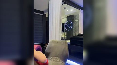 Media: Video of a modern, dimly-lit hotel room featuring a plush, grey armchair, a bed with black bedding, and a glass door with a Chanel logo.