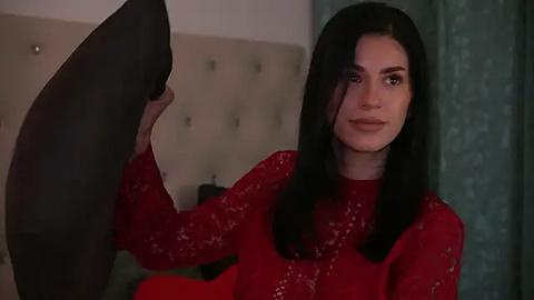 Media: Video of a woman with long black hair, fair skin, and wearing a red lace top, holding a black pillow, in a beige tufted headboard bedroom.