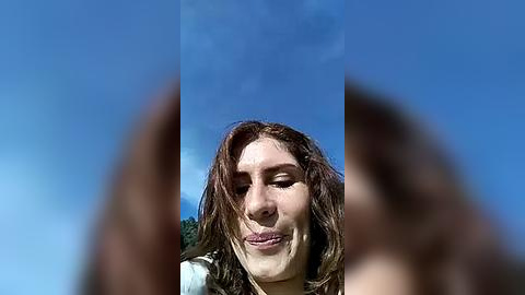 Media: Video of a woman with long brown hair, making a silly face, with her fingers in front, against a clear blue sky.