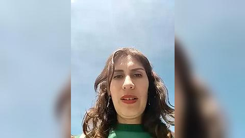 Media: Video of a young woman with long brown hair, wearing a green sweater, standing against a bright blue sky with sun rays filtering through clouds.